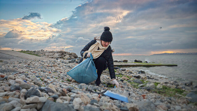 Young woman volunteer in action, wearing gloves and collecting trash on the shore, emphasizing the importance of maintaining clean and healthy oceans.