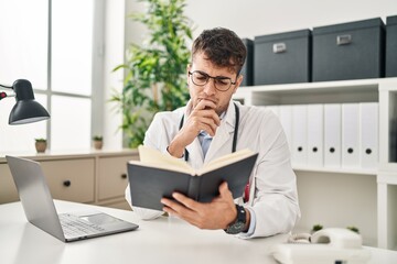 Young hispanic man doctor using laptop reading book at clinic