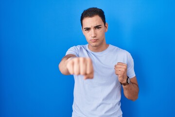 Young hispanic man standing over blue background punching fist to fight, aggressive and angry...