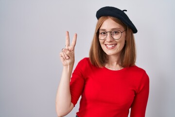 Young redhead woman standing wearing glasses and beret showing and pointing up with fingers number two while smiling confident and happy.