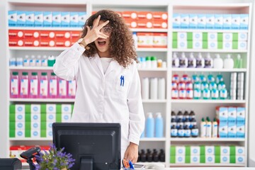 Hispanic woman with curly hair working at pharmacy drugstore peeking in shock covering face and eyes with hand, looking through fingers with embarrassed expression.