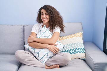 Hispanic woman with curly hair sitting on the sofa at home winking looking at the camera with sexy...