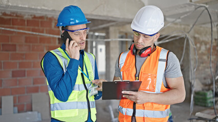 Two men builders writing document talking on smartphone at construction site