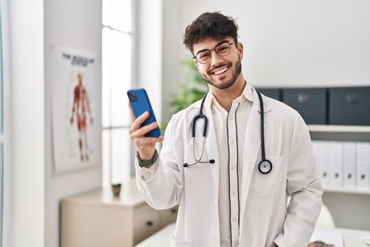 Young hispanic man wearing doctor uniform using smartphone at clinic - Powered by Adobe