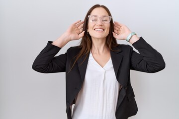 Beautiful brunette woman wearing business jacket and glasses smiling pulling ears with fingers,...