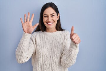 Young brunette woman standing over blue background showing and pointing up with fingers number six...