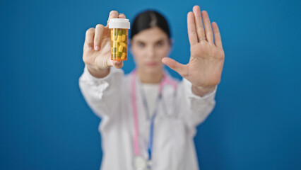 Young beautiful hispanic woman doctor holding pills doing stop gesture over isolated blue background