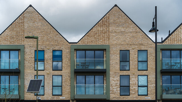 Symmetrical geometric rhythms on brick wall, triangular roofs, rectangular windows and balconies. Folding furniture on balconies. Two street lamps, solar panel in foreground. Cloudy English sky. - Powered by Adobe