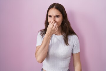 Young hispanic girl standing over pink background smelling something stinky and disgusting, intolerable smell, holding breath with fingers on nose. bad smell