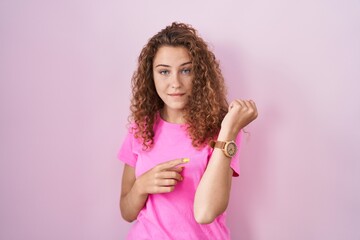 Young caucasian woman standing over pink background in hurry pointing to watch time, impatience,...