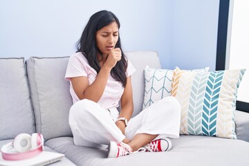 Young hispanic woman sitting on the sofa at home feeling unwell and coughing as symptom for cold or...
