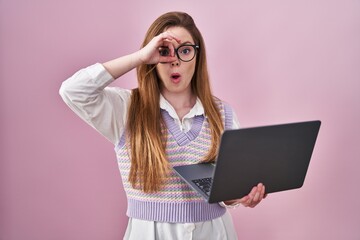 Young caucasian woman working using computer laptop doing ok gesture shocked with surprised face, eye looking through fingers. unbelieving expression.