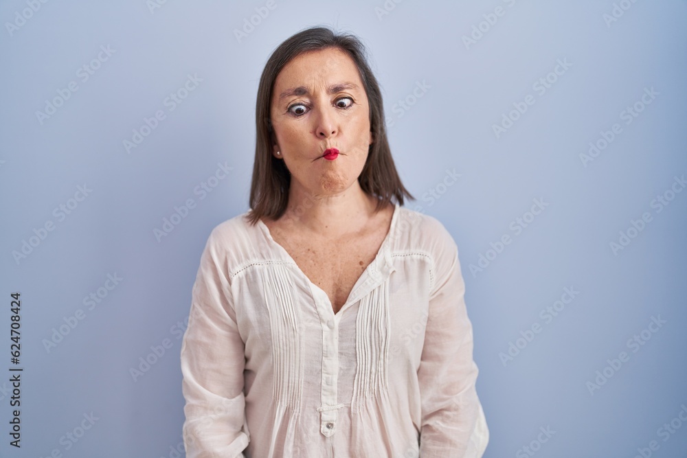 Wall mural middle age hispanic woman standing over blue background making fish face with lips, crazy and comica