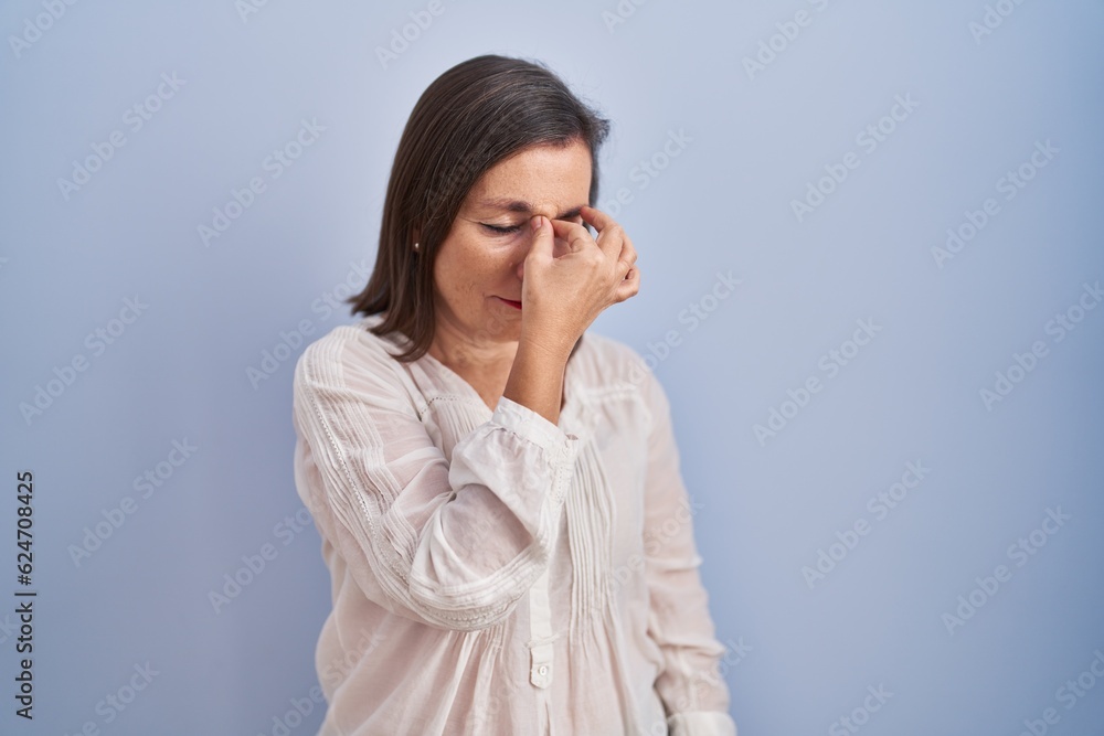 Poster Middle age hispanic woman standing over blue background tired rubbing nose and eyes feeling fatigue and headache. stress and frustration concept.