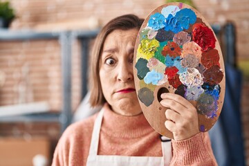 Middle age hispanic woman holding painter palette close to face skeptic and nervous, frowning upset because of problem. negative person.