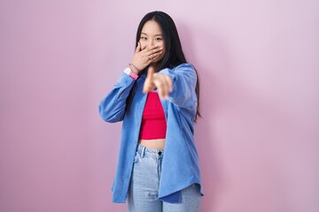 Young asian woman standing over pink background laughing at you, pointing finger to the camera with...