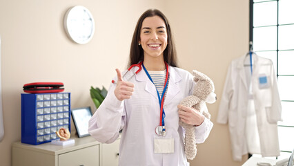 Young beautiful hispanic woman doctor holding teddy bear doing ok gesture at clinic