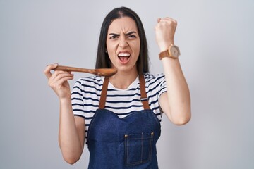 Young brunette woman wearing apron tasting food holding wooden spoon annoyed and frustrated...