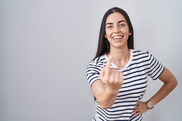 Young brunette woman wearing striped t shirt beckoning come here gesture with hand inviting welcoming happy and smiling