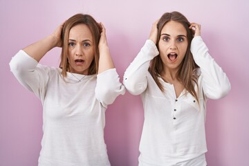 Middle age mother and young daughter standing over pink background crazy and scared with hands on head, afraid and surprised of shock with open mouth