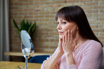 Middle aged beautiful woman looking at her face in mirror