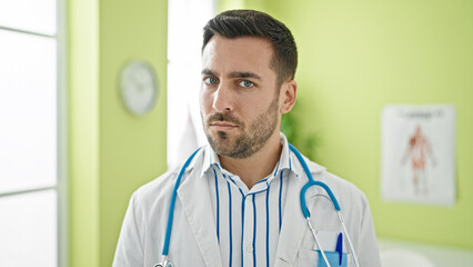 Young hispanic man doctor standing with serious expression at clinic