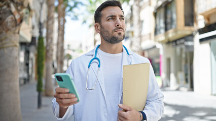 Young hispanic man doctor holding medical report using smartphone at street