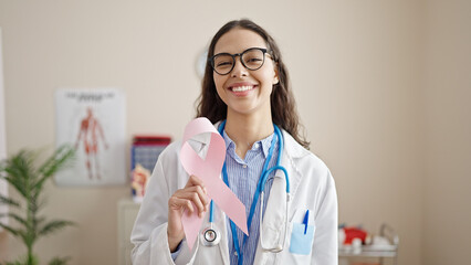 Young beautiful hispanic woman doctor holding breast cancer awareness pink ribbon at clinic