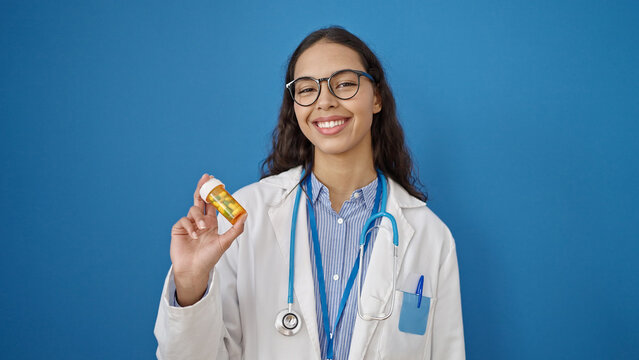 Young beautiful hispanic woman doctor holding pills over isolated blue background