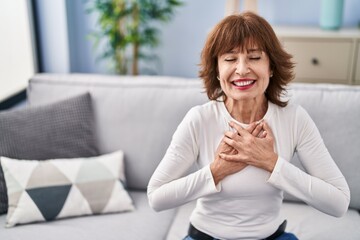 Middle age woman sitting on sofa with hands on heart at home