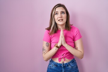 Blonde caucasian woman standing over pink background begging and praying with hands together with...