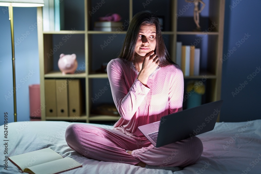 Sticker Young hispanic woman using computer laptop on the bed with hand on chin thinking about question, pensive expression. smiling with thoughtful face. doubt concept.