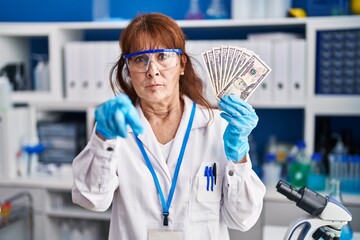Middle age hispanic woman working at scientist laboratory holding dollars pointing with finger to the camera and to you, confident gesture looking serious