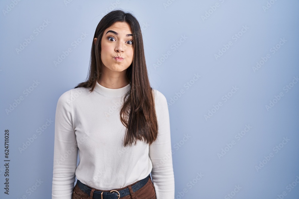 Sticker Young brunette woman standing over blue background puffing cheeks with funny face. mouth inflated with air, crazy expression.