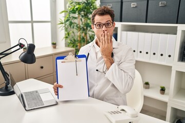 Young hispanic man wearing doctor stethoscope holding clipboard covering mouth with hand, shocked and afraid for mistake. surprised expression