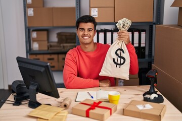Hispanic man working at small business ecommerce holding money bag looking positive and happy...