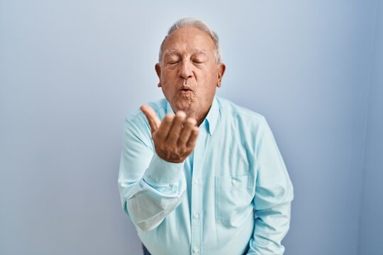 Senior Man With Grey Hair Standing Over Blue Background Looking At The Camera Blowing A Kiss With Hand On Air Being Lovely And Sexy. Love Expression.