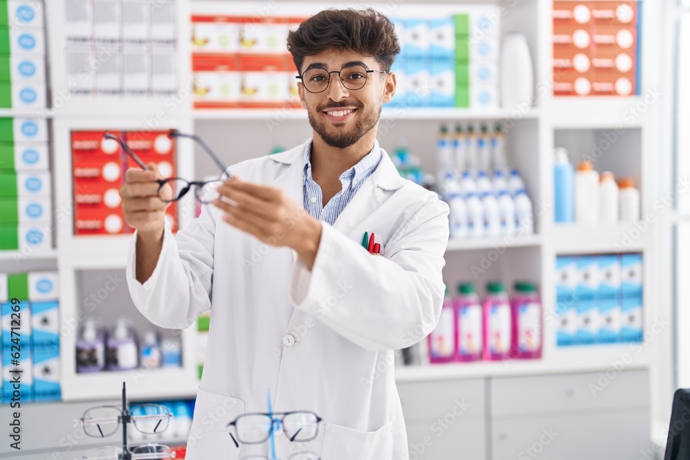 Sticker young arab man pharmacist smiling confident holding glasses at pharmacy