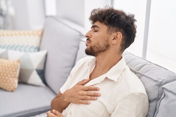 Young arab man suffering heart attack sitting on sofa at home