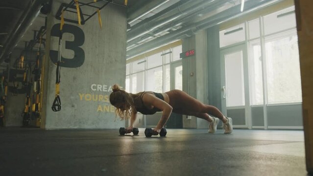 Beautiful strong woman doing push-ups using dumbbells alone in the gym studio
