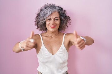 Middle age woman with grey hair standing over pink background approving doing positive gesture with hand, thumbs up smiling and happy for success. winner gesture.