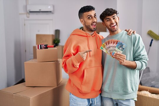 Young hispanic gay couple moving to a new home holding banknotes smiling happy pointing with hand and finger to the side