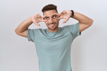 Hispanic man with beard standing over white background doing peace symbol with fingers over face, smiling cheerful showing victory