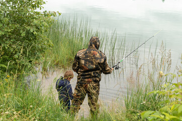 Side view portrait of father and son sitting together on rocks fishing with rods in calm lake waters with landscape of setting sun, both wearing checkered shirts, shot from behind tree