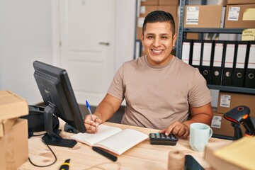 Young latin man ecommerce business worker using calculator writing on notebook at office