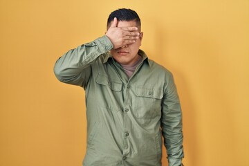 Hispanic young man standing over yellow background covering eyes with hand, looking serious and...