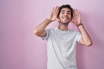 Young hispanic man standing over pink background smiling cheerful playing peek a boo with hands...
