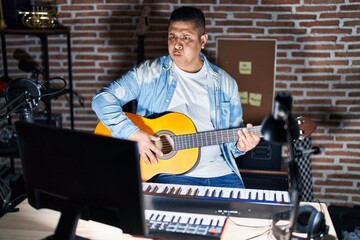 Hispanic young man playing classic guitar at music studio puffing cheeks with funny face. mouth inflated with air, crazy expression.