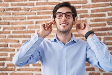 Young hispanic man standing over brick wall background smiling pulling ears with fingers, funny...
