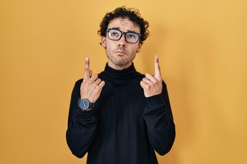 Hispanic man standing over yellow background pointing up looking sad and upset, indicating direction with fingers, unhappy and depressed.